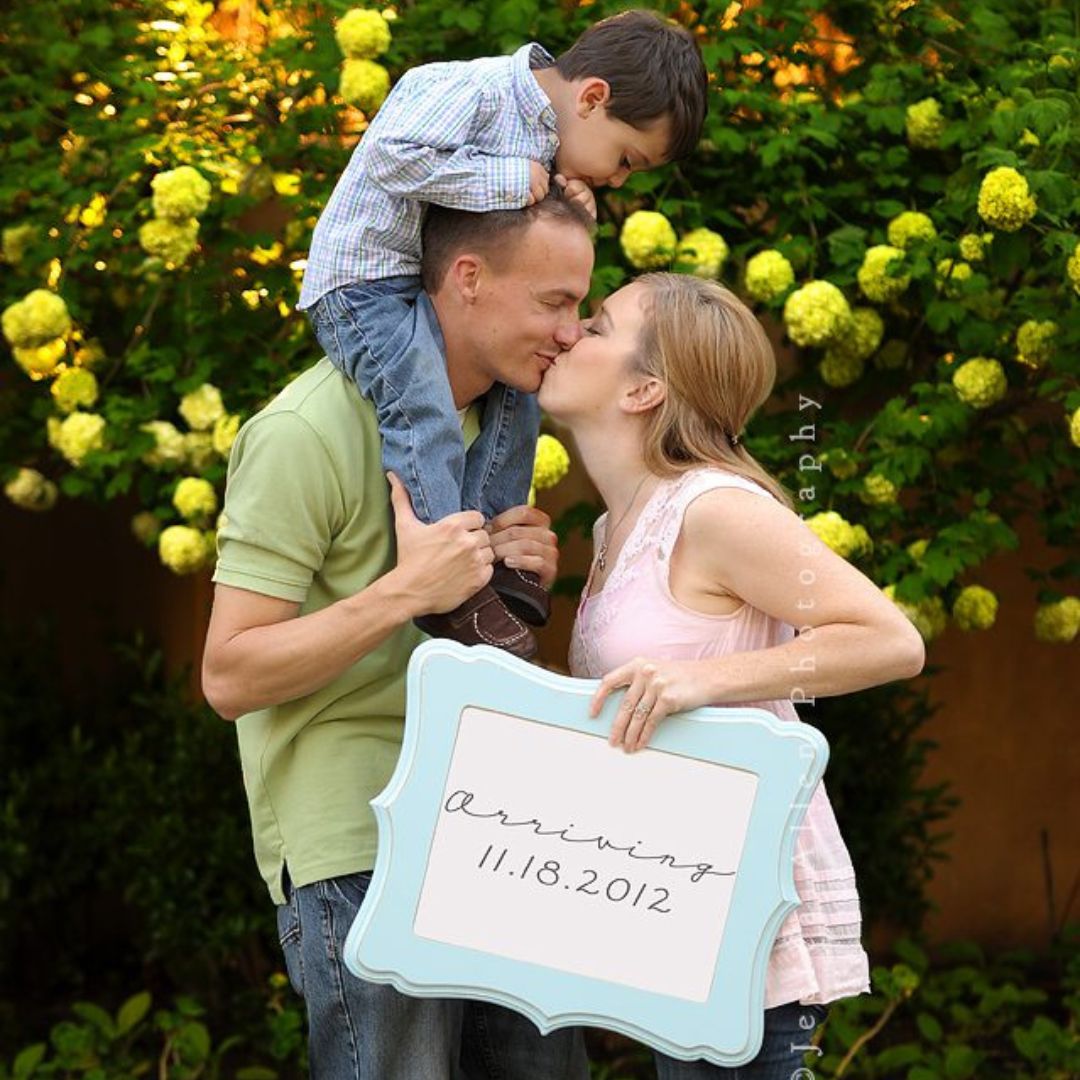 a family posing happily kissing with their son while holding a Baby number two sign