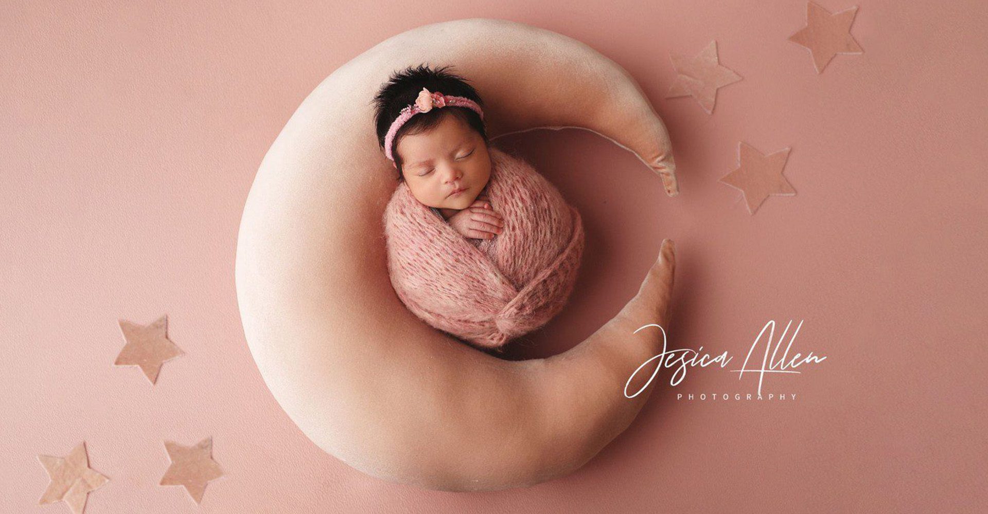 A photoshoot with a newborn girl in pink laying in the middle of a pink crescent shaped pillow